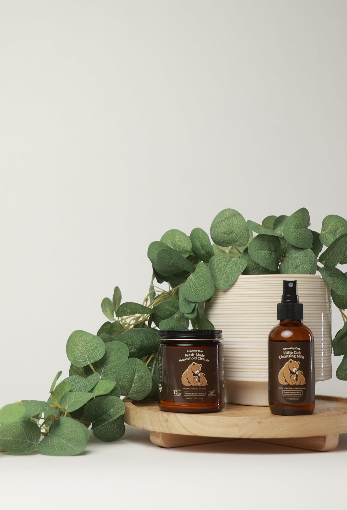 Two amber glass bottles and a matching amber jar with cream labels, styled on a round wooden tray with green eucalyptus leaves and a white ceramic pot against a light gray background.