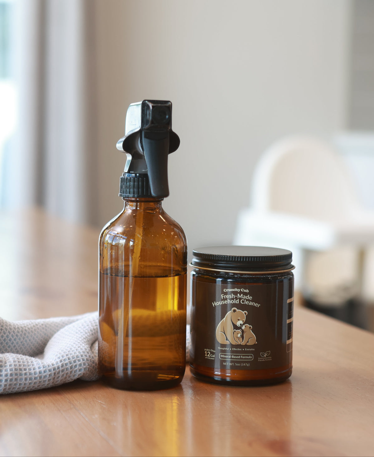 An amber glass spray bottle and a matching amber jar with a black lid sit on a wooden table in a softly blurred indoor setting. The jar label shows a dog and cat illustration with the brand name 'Pup & Cat' visible.