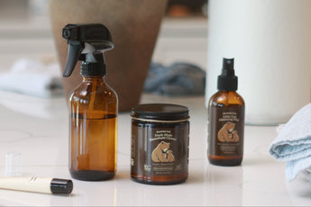Three amber glass home fragrance products, including a spray bottle, a jar, and a smaller pump bottle, arranged on a white countertop in a kitchen setting with a vase of white flowers in the background.
